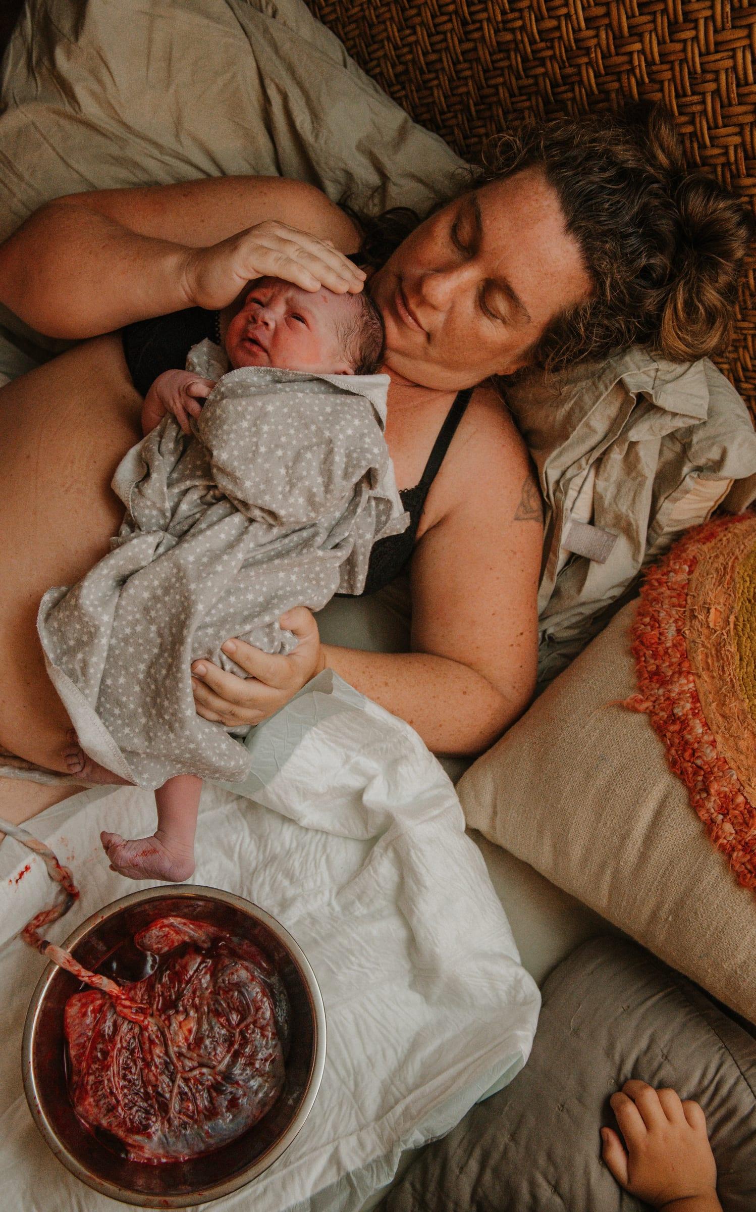 A mother resting with her eyes closed, cradling her newborn on her chest just after birth, the placenta in a bowl beside them